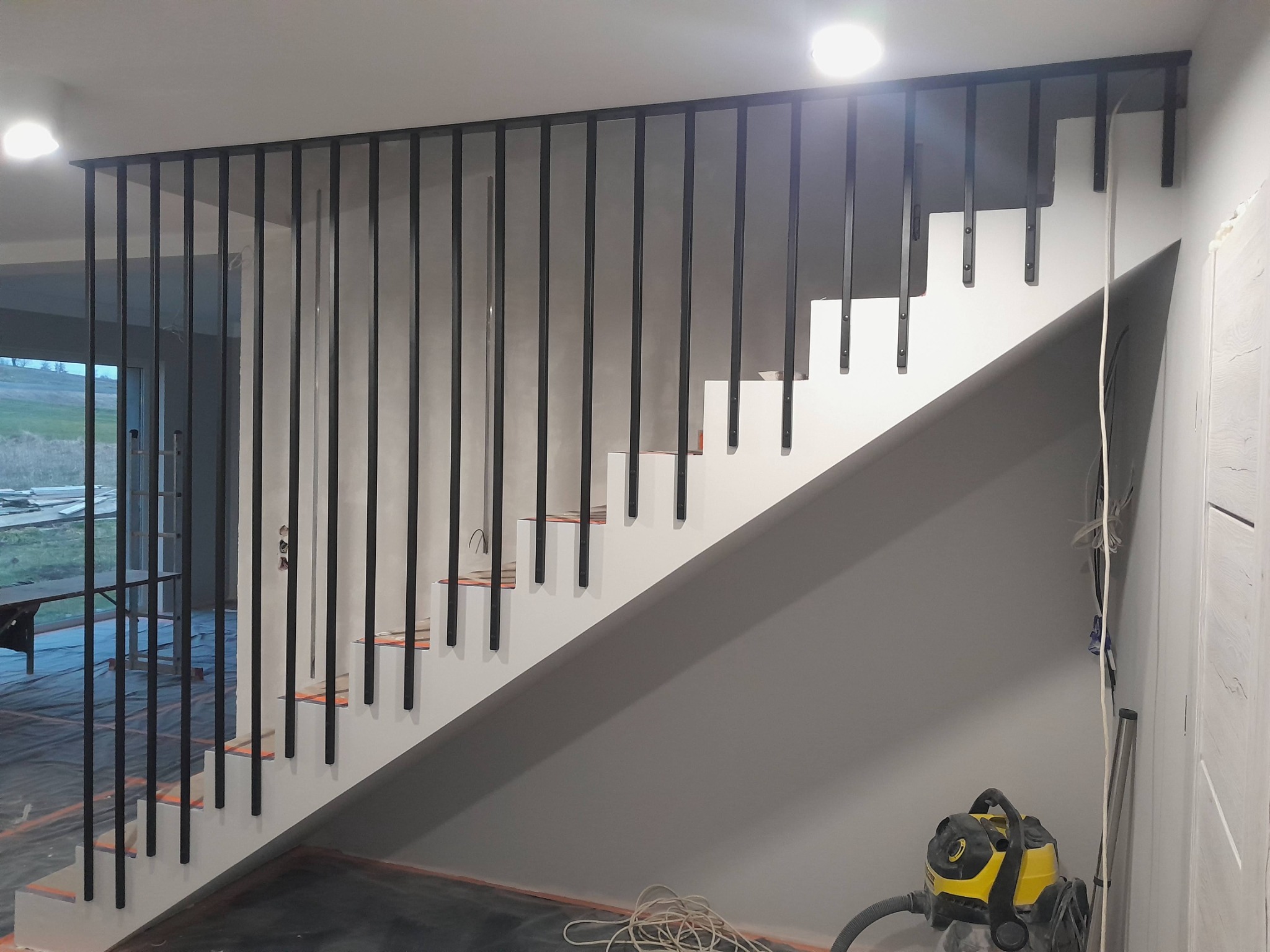 Nowoczesne schody z czarną metalową balustradą w Żukowie, widoczne podczas montażu, z odkurzaczem obok ściany.