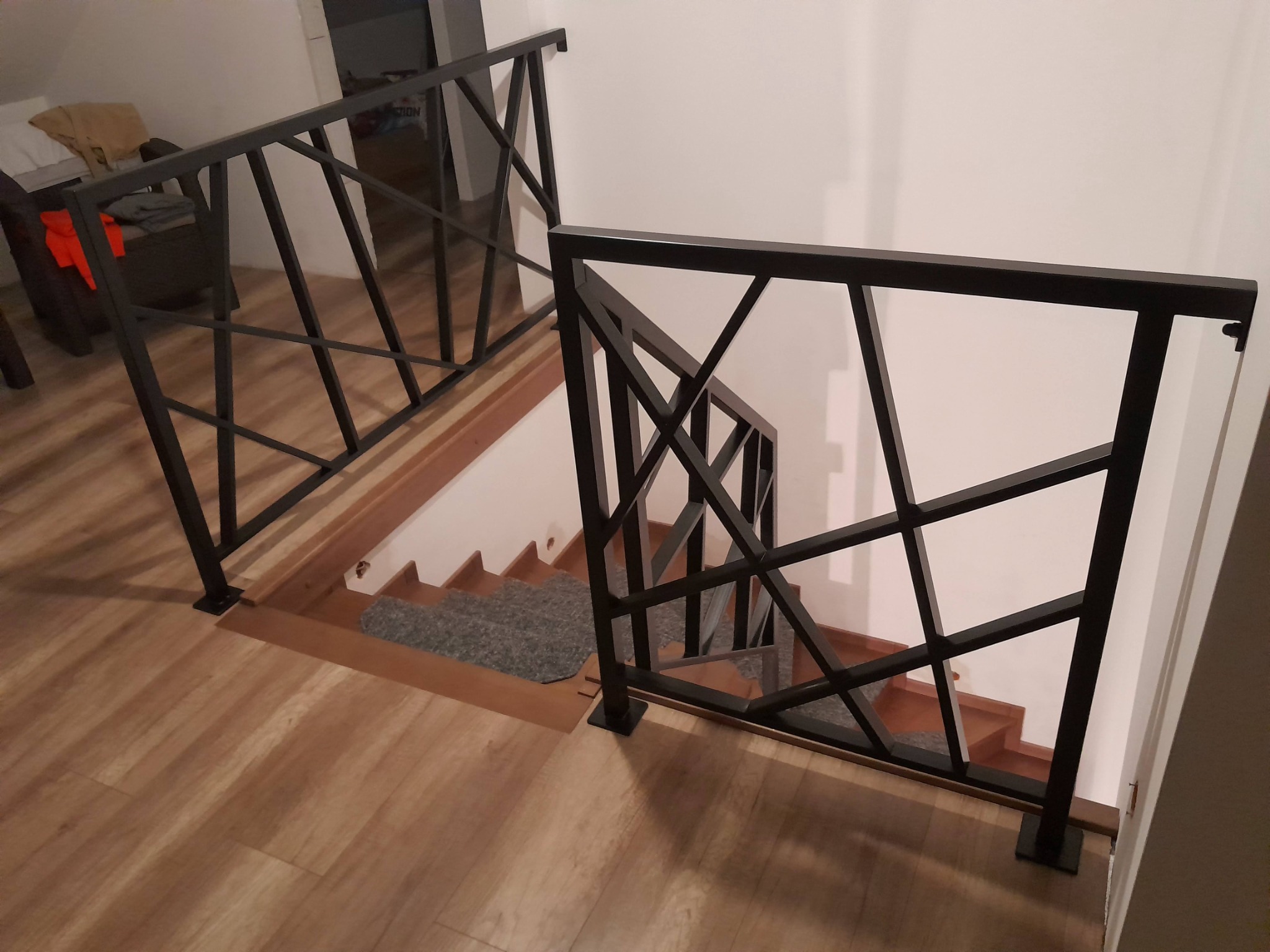 Klatka schodowa z nowoczesną, geometryczną balustradą w kolorze czarnym, wykonaną z metalowych profili, widok z góry na drewniane schody i podłogę.