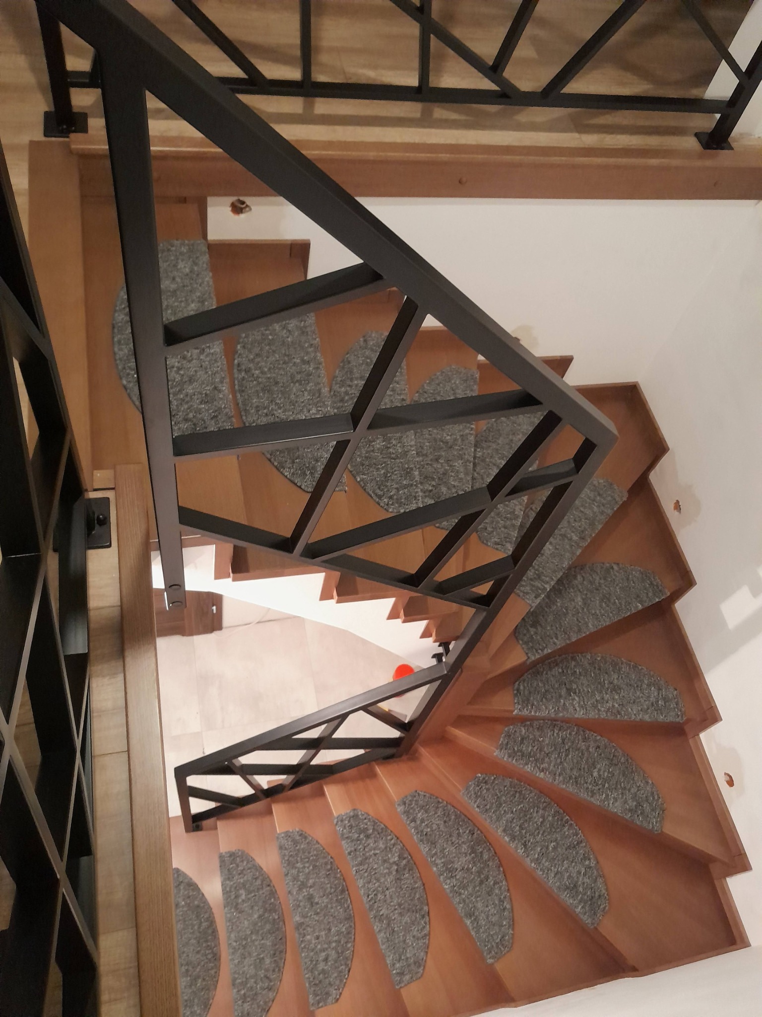Wysoki kąt widzenia na drewniane schody spiralne z szarymi nakładkami dywanowymi i nowoczesną, geometryczną, czarną metalową balustradą.