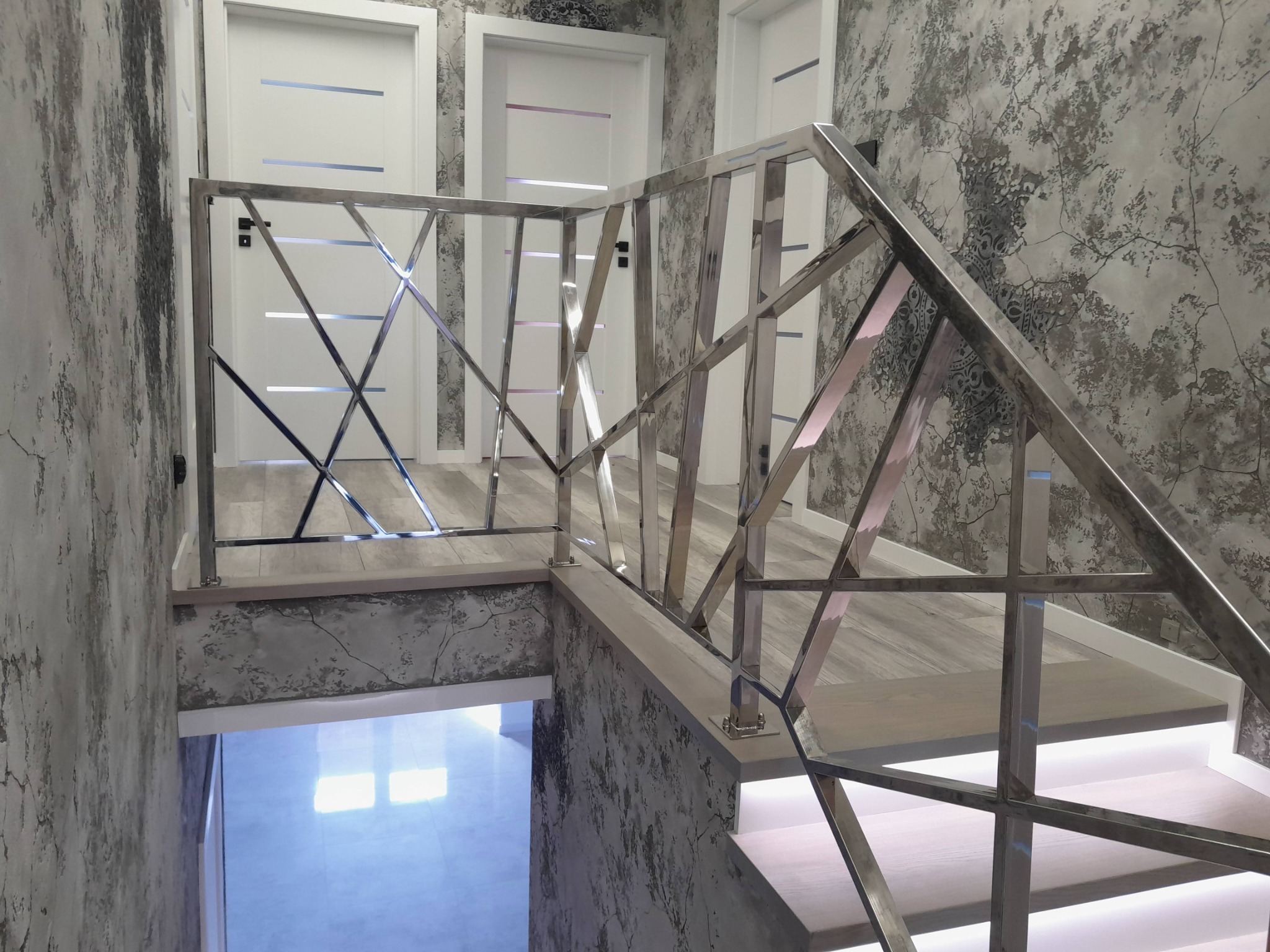 Nowoczesna, metalowa balustrada schodowa z geometrycznym wzorem, polerowana na wysoki połysk, na tle ściany o wzorzystej strukturze i oświetlonych schodów.