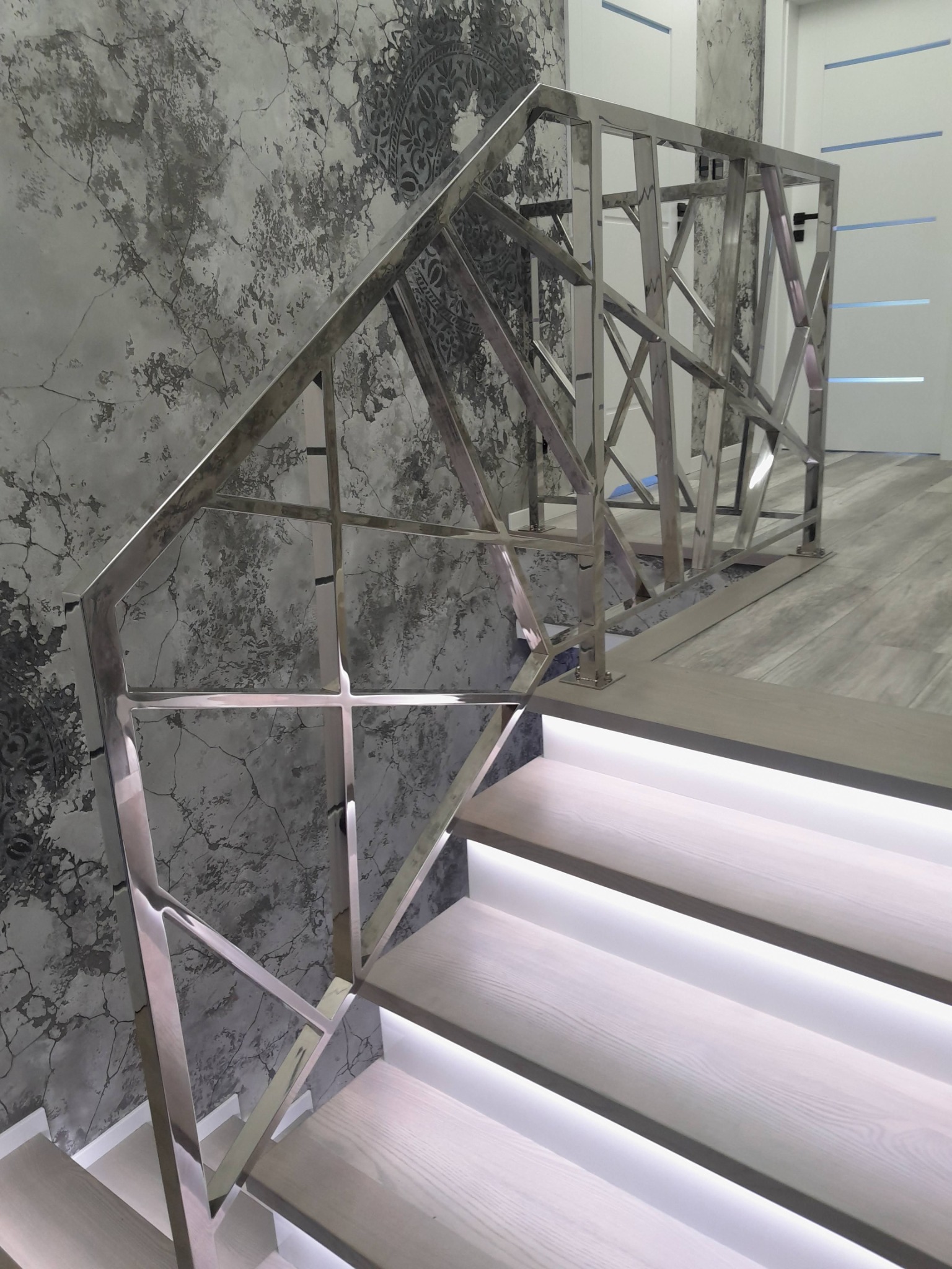 Nowoczesna balustrada nierdzewna o geometrycznym wzorze na schodach z podświetleniem LED, na tle ściany z efektem betonu.