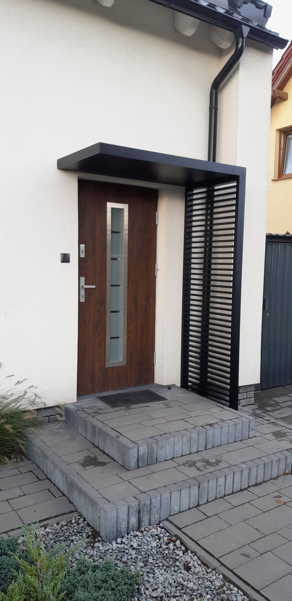 Nowoczesne zadaszenie nad wejściem do domu, z metalową konstrukcją w kolorze czarnym i drewnianymi drzwiami z elementami szkła. Schody wejściowe wyłożone szarą kostką.