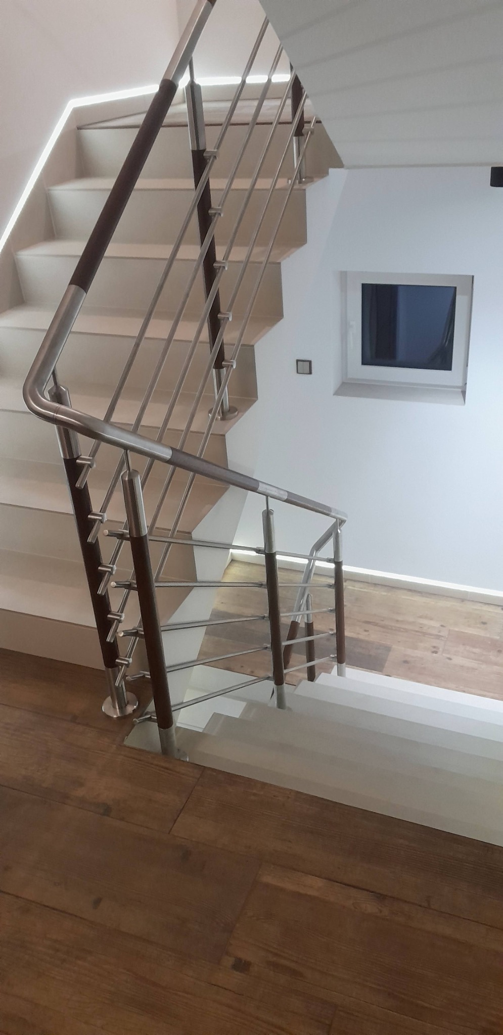 Nowoczesne schody z metalową balustradą i drewnianymi poręczami, oświetlone listwą LED. Widok z góry na drewnianą podłogę, białe stopnie i okno w tle.