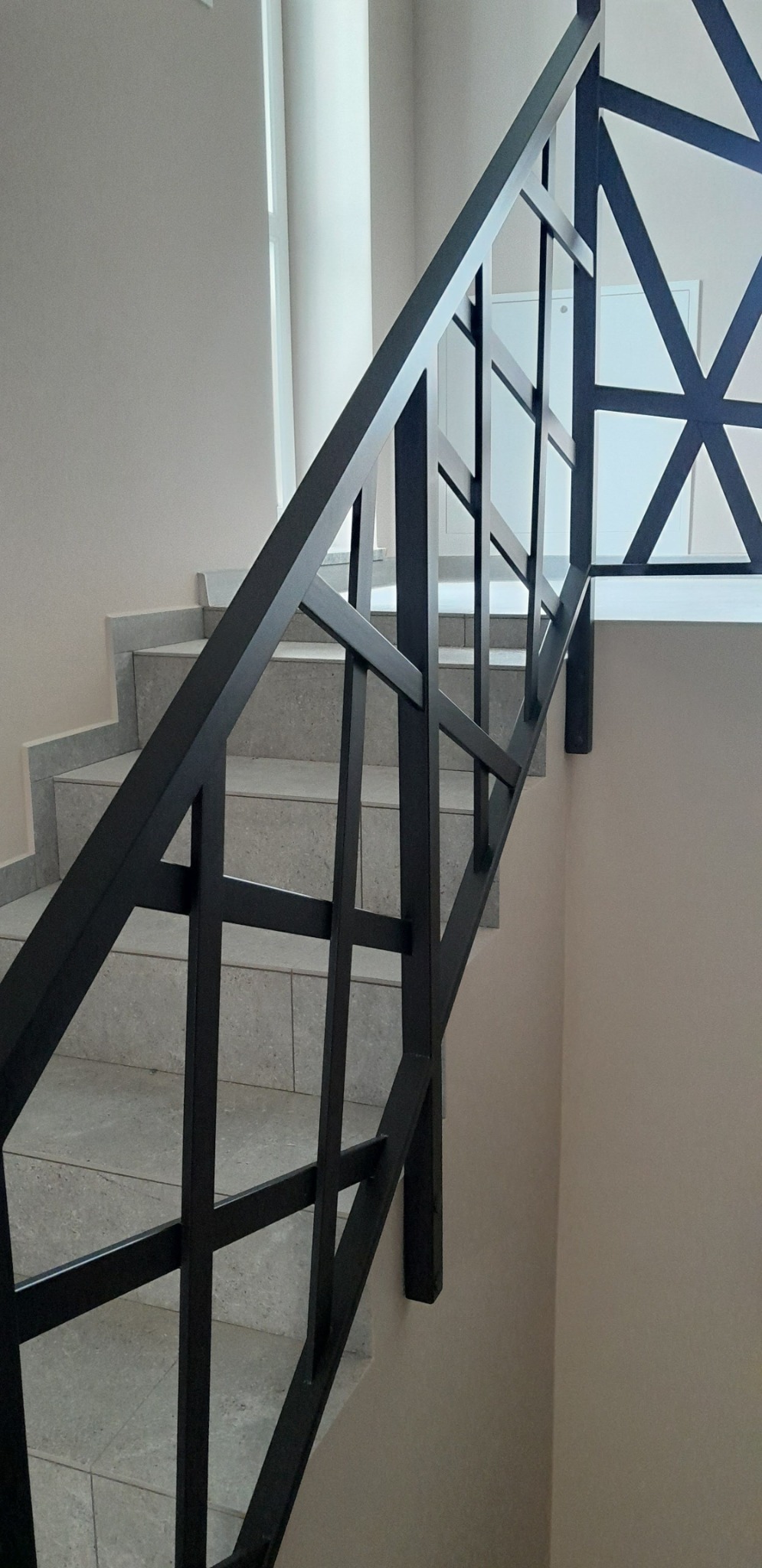 Nowoczesna, czarna poręcz schodowa z geometrycznym wzorem, zamontowana w jasnym wnętrzu. Widoczne szare stopnie schodów i fragment białej ściany.