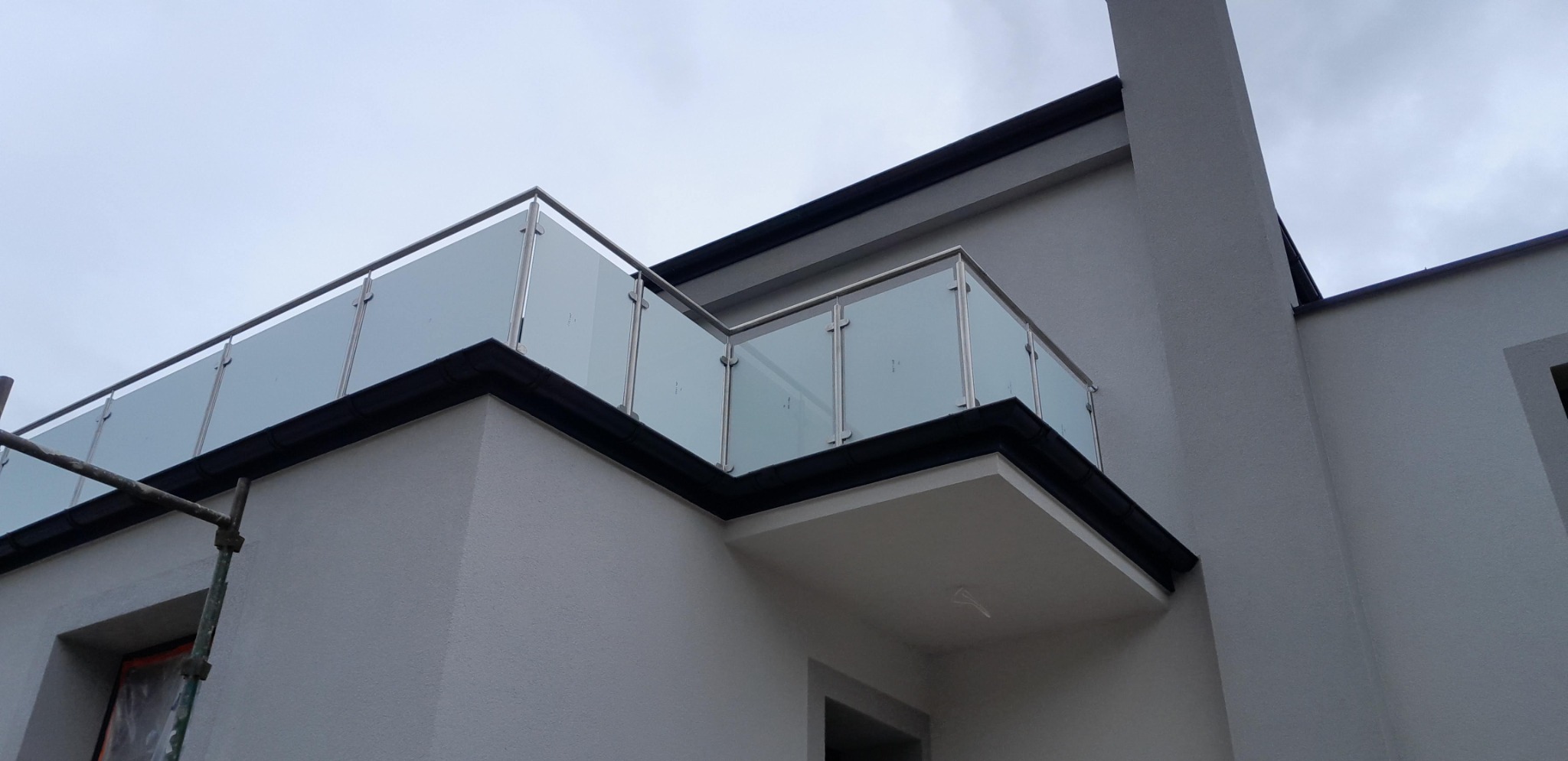 Szklana balustrada z nierdzewnymi elementami na balkonie budynku o szarej elewacji, widok z dołu, fragment rusztowania z lewej strony kadru.