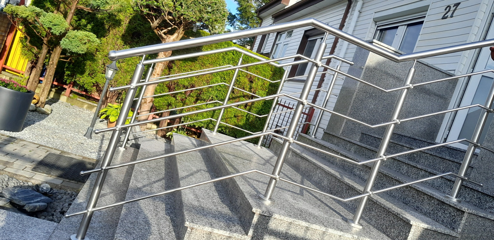 Nowoczesna balustrada ze stali nierdzewnej na granitowych schodach wejściowych do domu z numerem 27, otoczona zielenią.