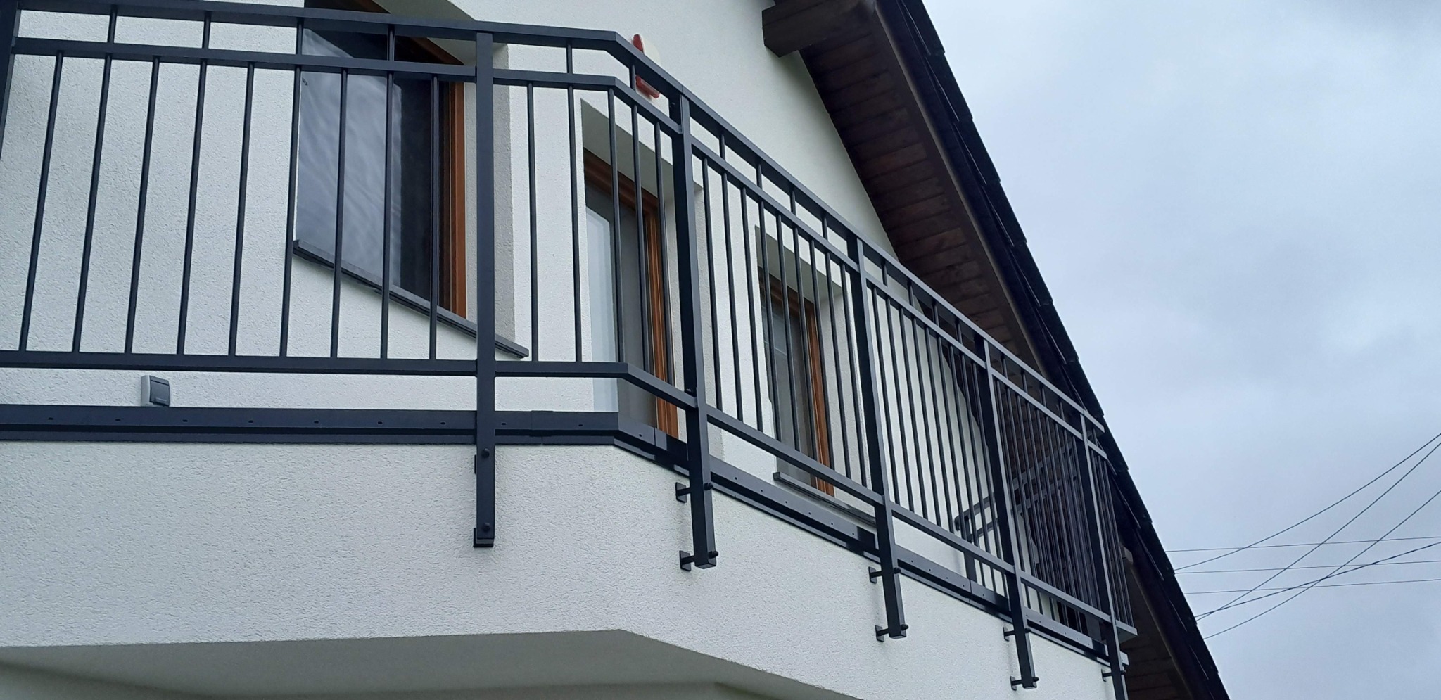 Czarna, metalowa balustrada balkonowa z pionowymi szczebelkami, zamontowana na białej elewacji domu. Widoczne okna i fragment dachu. Ujęcie z dołu, pochmurne niebo.