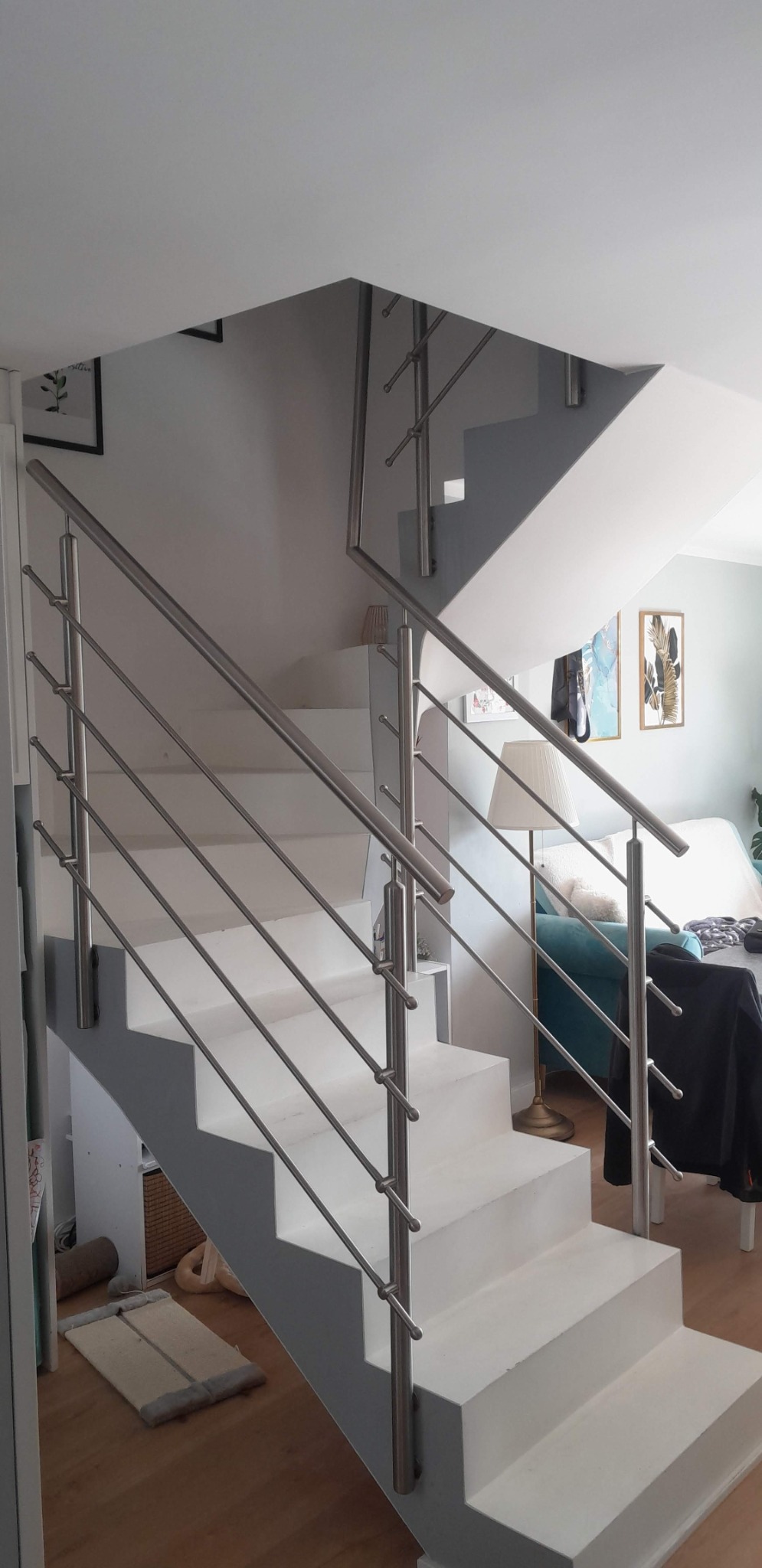 Wewnętrzne schody z białymi stopniami i nowoczesną, metalową balustradą z poziomymi prętami. W tle widoczny salon z turkusową sofą i lampą podłogową.