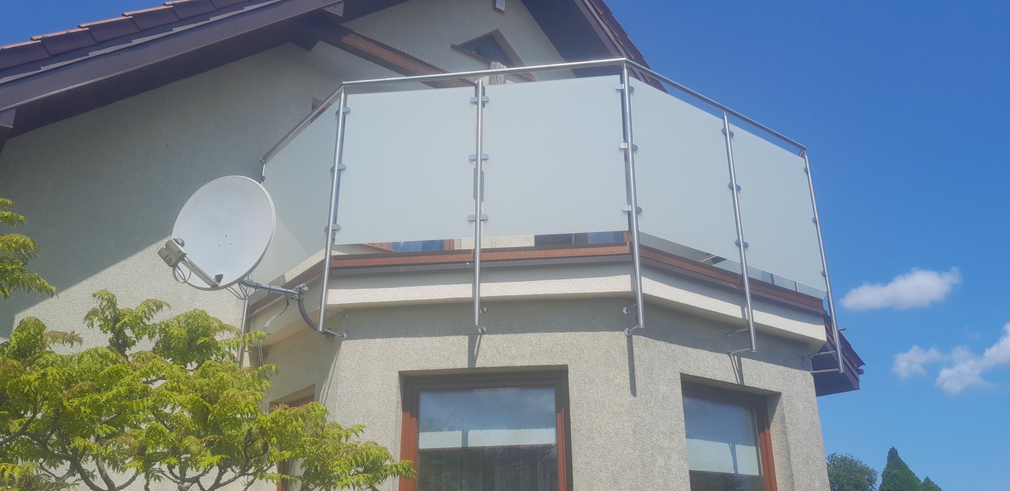 Szklana balustrada balkonowa z nierdzewnymi słupkami na tle błękitnego nieba, widoczna antena satelitarna i fragment zieleni. Kąt ujęcia od dołu podkreśla nowoczesny design.