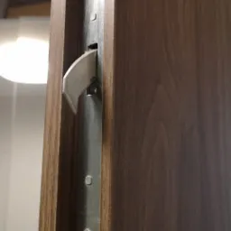 Zamek hakowy wpuszczany w brązowe drewniane drzwi przesuwne, widoczne śruby mocujące i mechanizm blokujący.