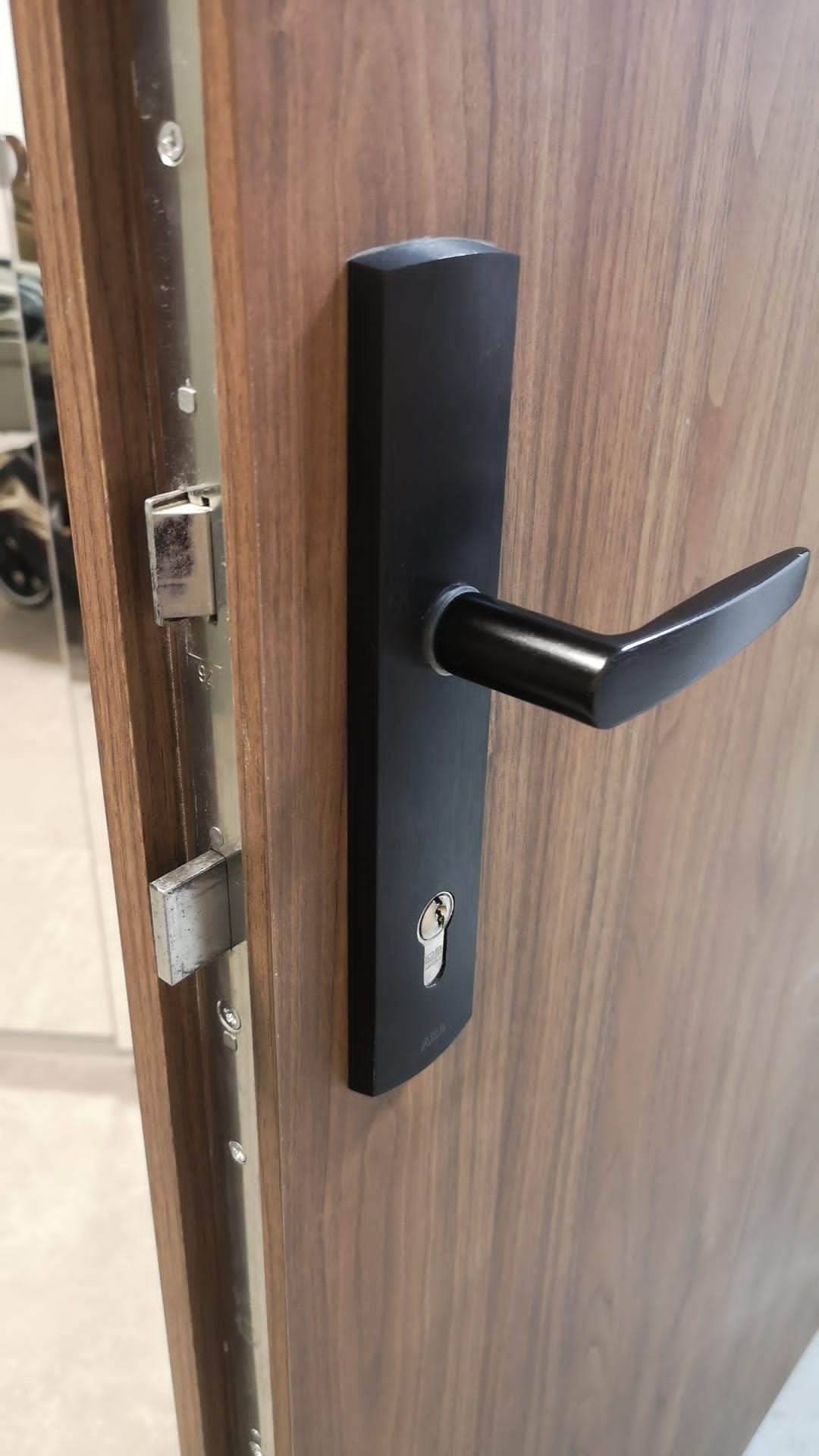 Zamknięte drzwi z drewnianą okleiną i czarną klamką z zamkiem, widoczny mechanizm zamka w ościeżnicy.