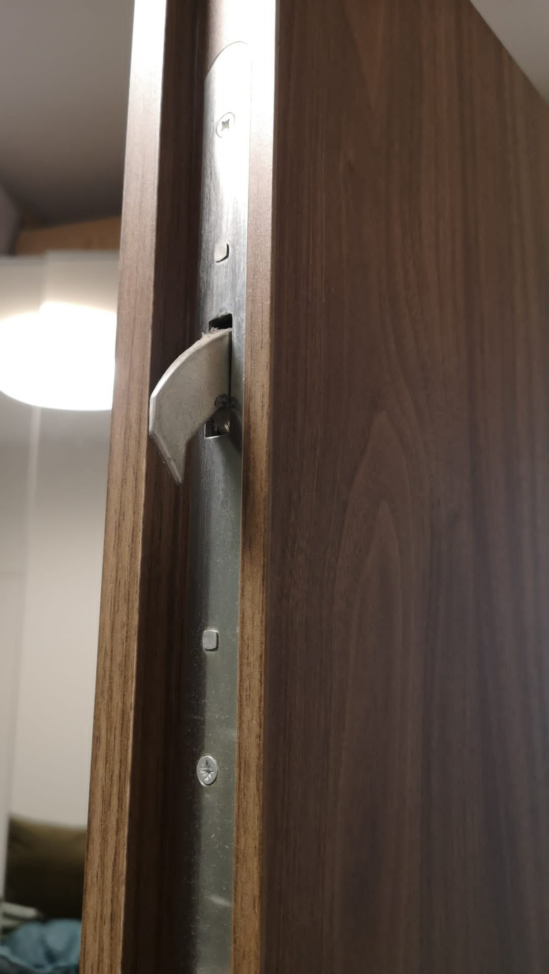 Zamek hakowy wpuszczany w brązowe drewniane drzwi przesuwne, widoczne śruby mocujące i mechanizm blokujący.