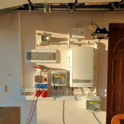 Instalacja elektryczna z inwerterem SolarEdge, zabezpieczeniami i gniazdami siłowymi zamontowana na ścianie garażu, widoczne przewody pomiarowe z miernikiem.