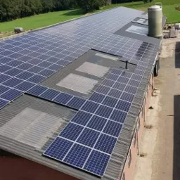 Rozległy dach budynku gospodarczego pokryty panelami słonecznymi, widoczny zbiornik na wodę, w tle zielone pola i las.