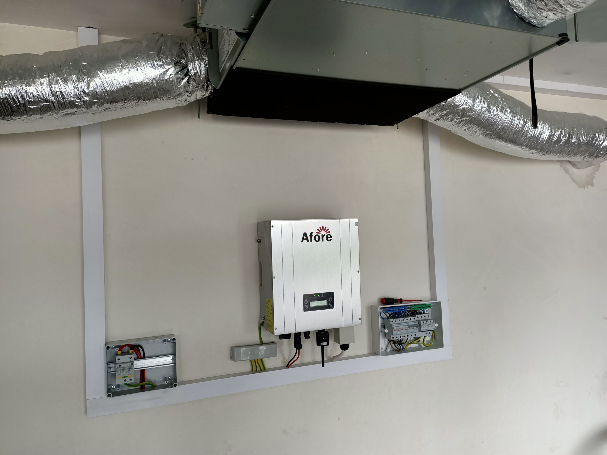 Zamontowany inwerter Afore na ścianie, obok skrzynki elektrycznej, z widocznymi przewodami i elementami instalacji, całość w ramie z białej listwy, pod sufitem z instalacją wentylacyjną.