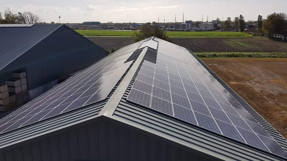 Panel słoneczny na dachu budynku gospodarczego z widokiem na pola uprawne i oddalony krajobraz przemysłowy.