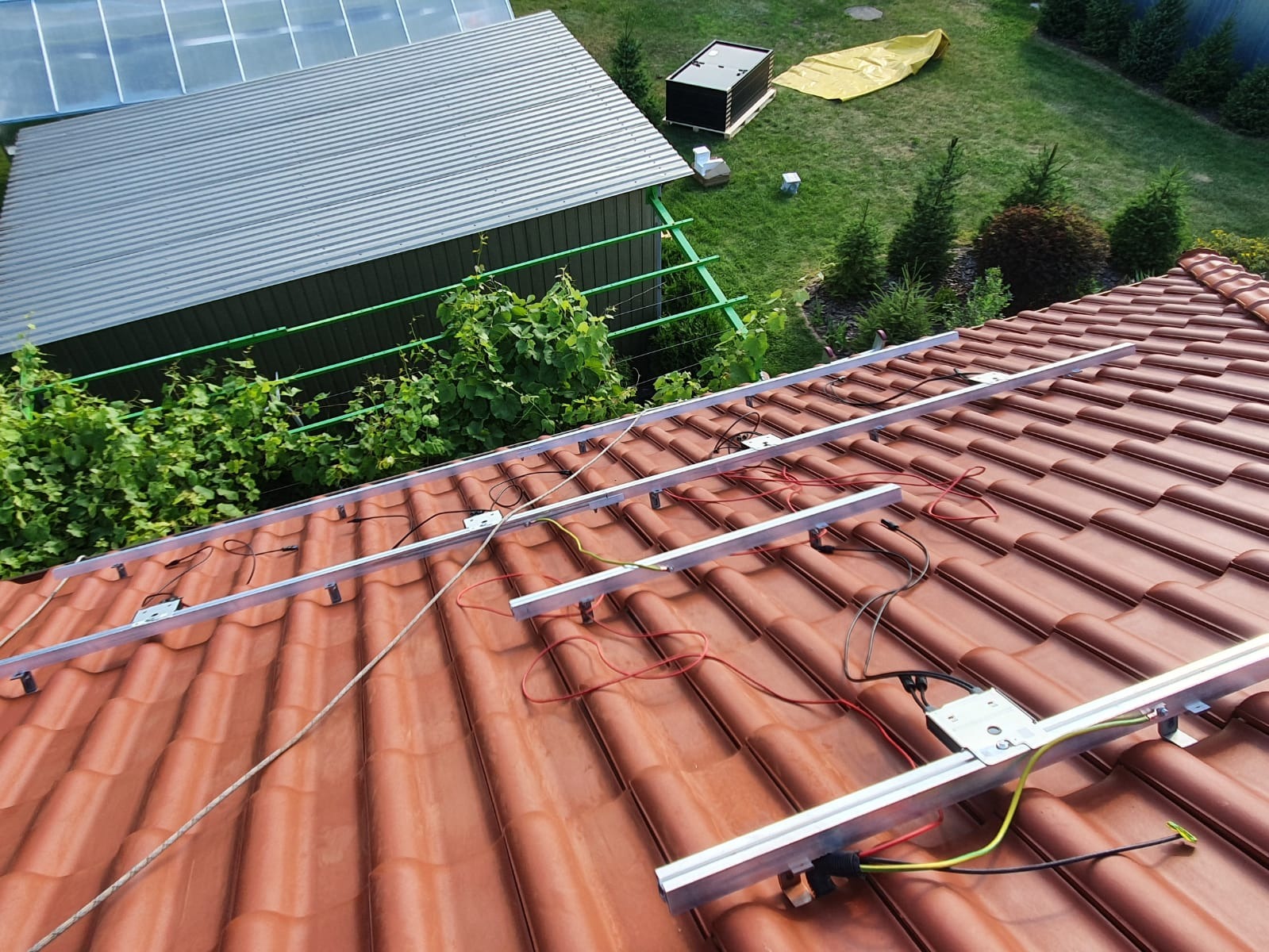 Montaż szyn pod panele słoneczne na dachu z czerwonej dachówki, widok z góry na częściowo zamontowaną instalację, z zielenią w tle.