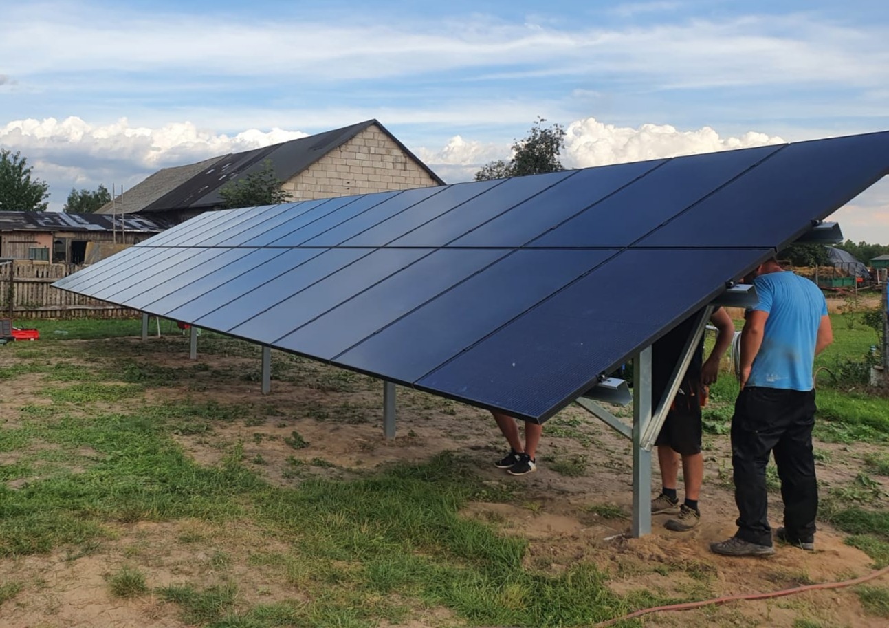 Montaż paneli słonecznych na konstrukcji wsporczej na trawiastym terenie, widoczni dwaj instalatorzy w trakcie pracy, w tle budynek gospodarczy i błękitne niebo z obłokami.