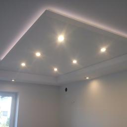 Usługi Remontowo-Wykończeniowe - Biały sufit podwieszany z oświetleniem LED w kształcie prostokąta i punktowymi lampami halogenowymi, widok z dołu na fragment pokoju z oknem.