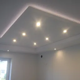 Biały sufit podwieszany z oświetleniem LED w kształcie prostokąta i punktowymi lampami halogenowymi, widok z dołu na fragment pokoju z oknem.