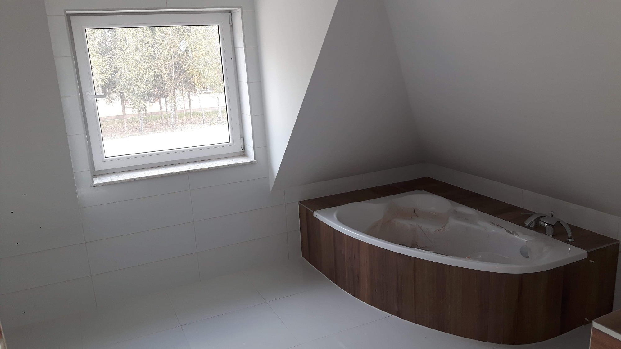 Nowoczesna łazienka na poddaszu z narożną wanną obudowaną drewnopodobną płytą i białymi płytkami, widok na okno z widokiem drzew
