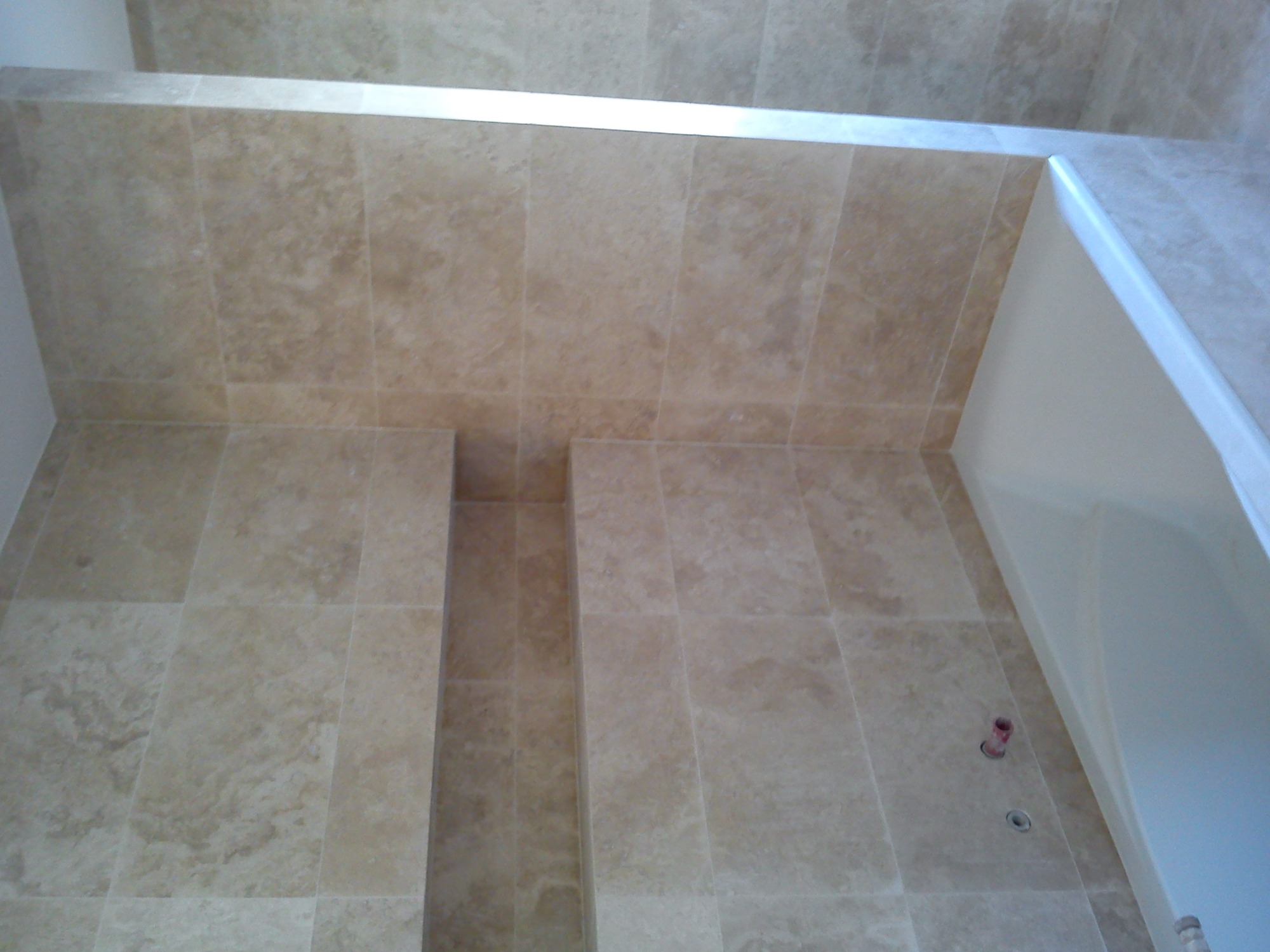 Wykończona ściana i sufit w łazience, pokryte beżowymi płytkami imitującymi trawertyn, z wnękami na kosmetyki i widocznymi przyłączami wodno-kanalizacyjnymi.