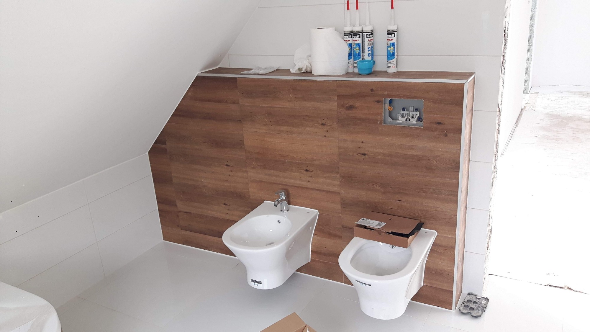 Bidet i toaleta w trakcie montażu w łazience na poddaszu, obudowa ze wzorem drewna, białe płytki i widoczne tuby silikonu.