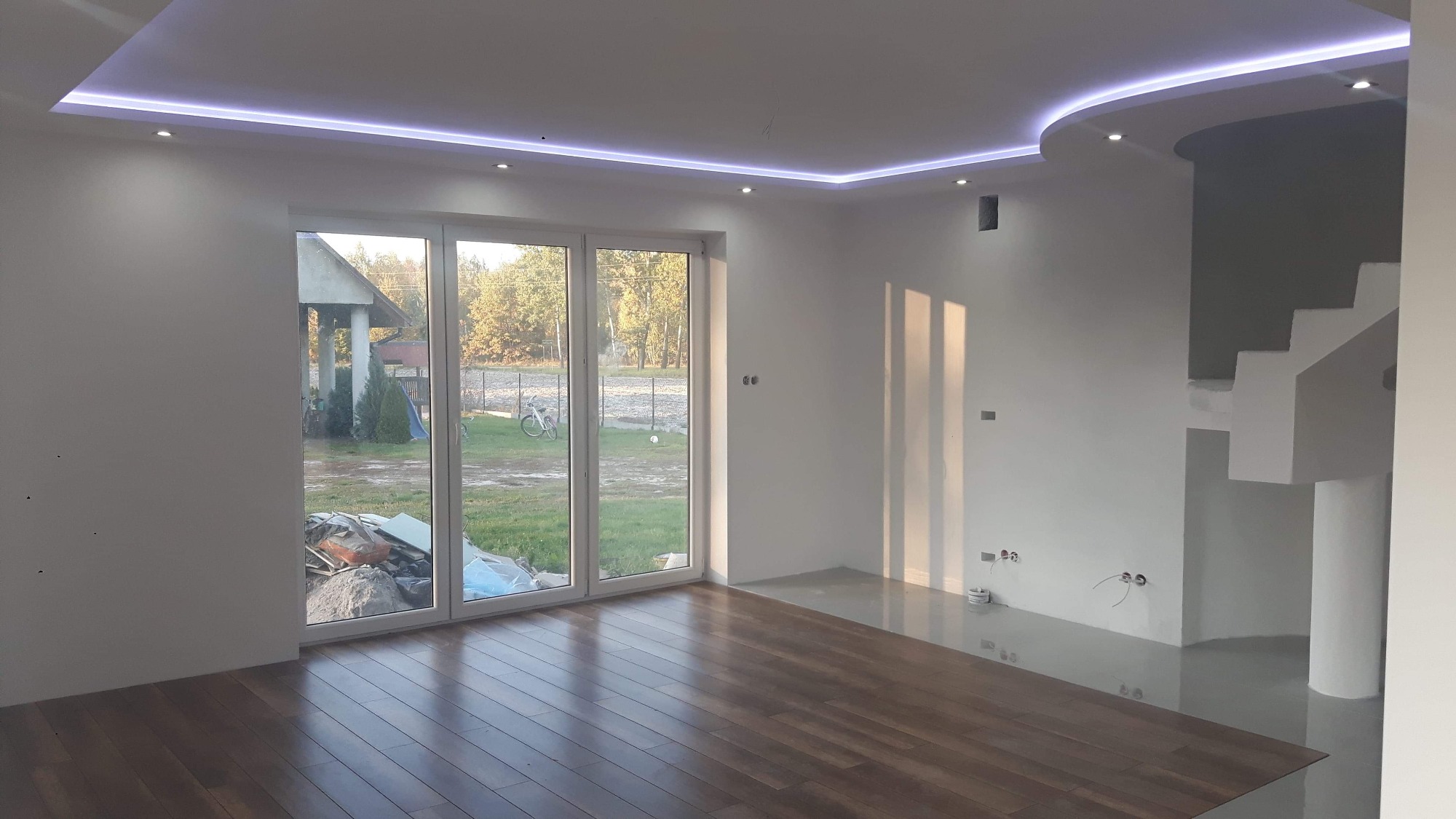 Salon po remoncie z drewnianą podłogą, białymi ścianami, oknem na ogród oraz podwieszanym sufitem z oświetleniem LED, częściowo wykończone schody w tle.