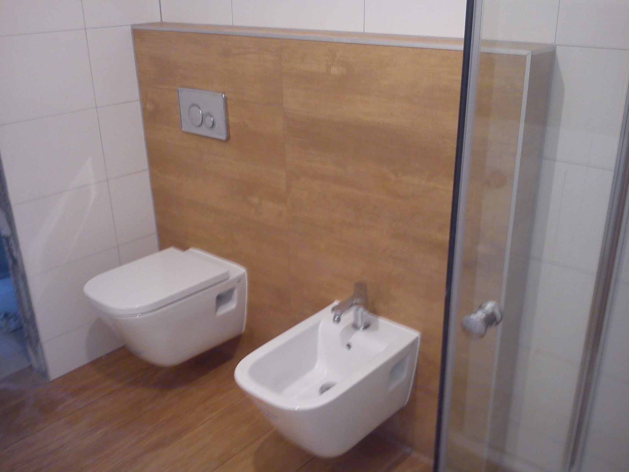 Nowoczesna łazienka z podwieszaną toaletą i bidetem, wykończona płytkami imitującymi drewno, z widoczną kabiną prysznicową z metalowym uchwytem.