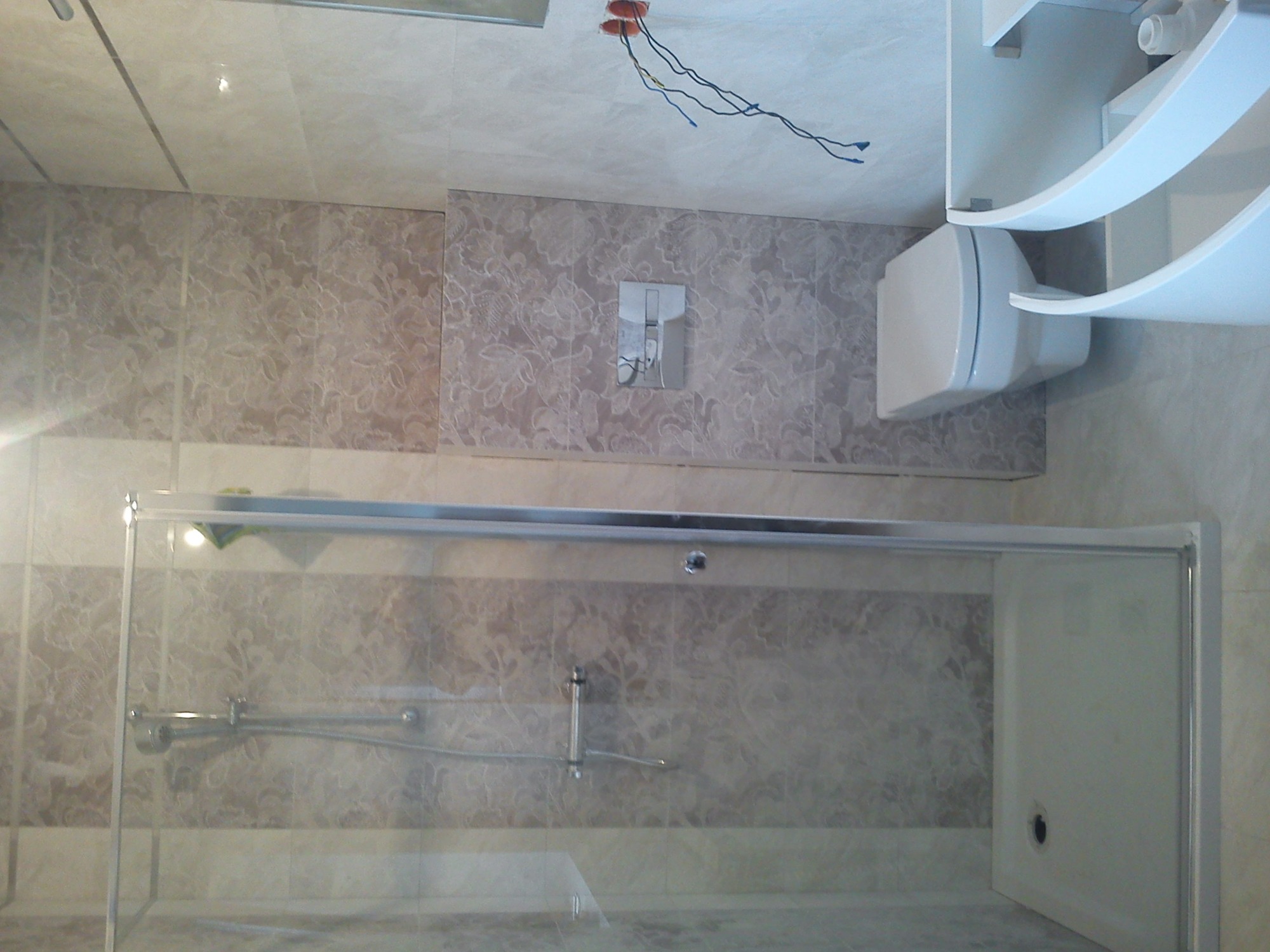 Wykończona łazienka z beżowymi płytkami z motywem kwiatowym, kabiną prysznicową z przesuwnymi drzwiami, wiszącą toaletą i odsłoniętymi przewodami elektrycznymi w suficie.