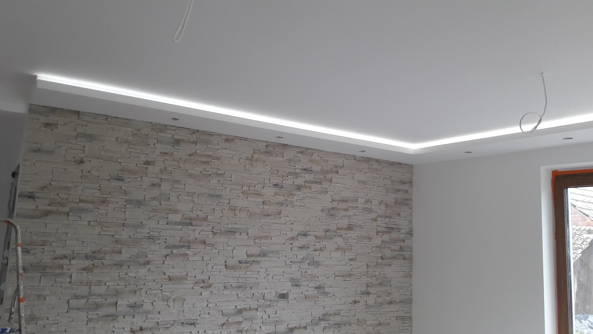 Ściana wykończona dekoracyjnym kamieniem z podświetleniem LED w suficie podwieszanym, widoczna drabina malarska w rogu pomieszczenia.