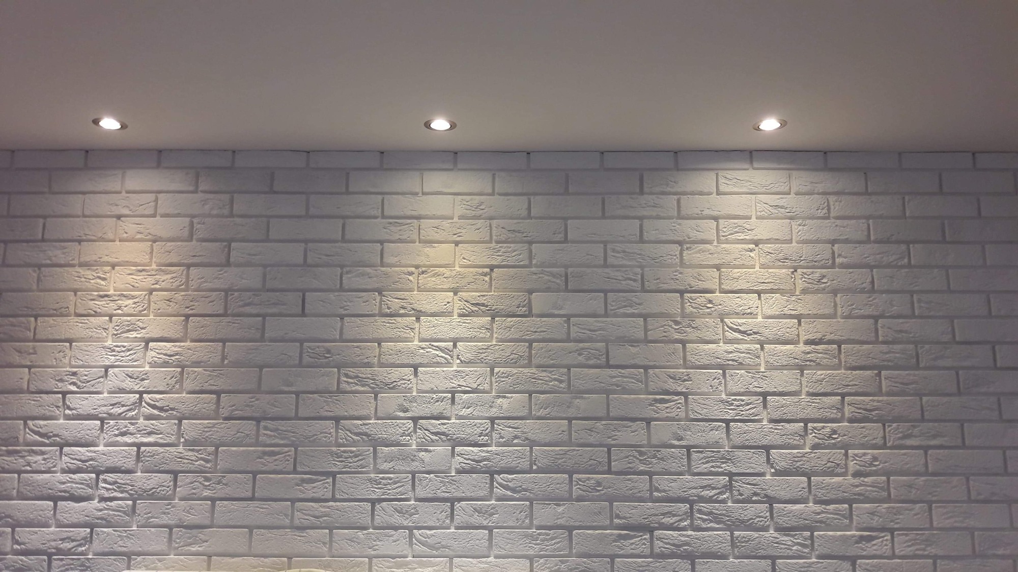 Ściana z białej cegły dekoracyjnej oświetlona trzema punktowymi lampami sufitowymi