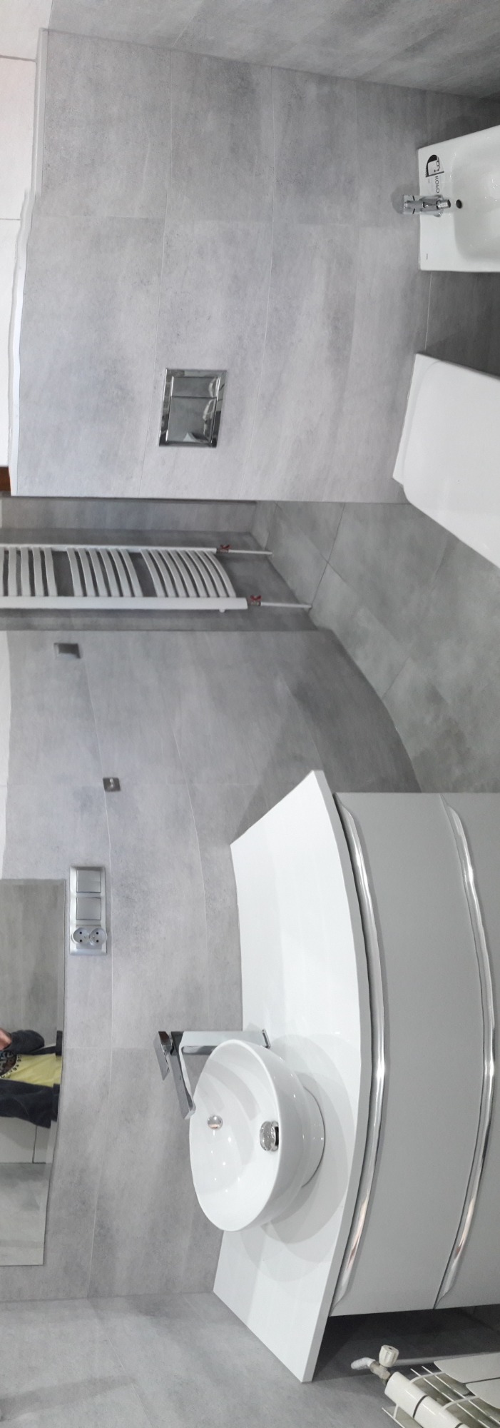 Nowoczesna łazienka z szarymi płytkami, umywalką na szafce z chromowanymi detalami, grzejnikiem drabinkowym i podtynkową spłuczką WC.