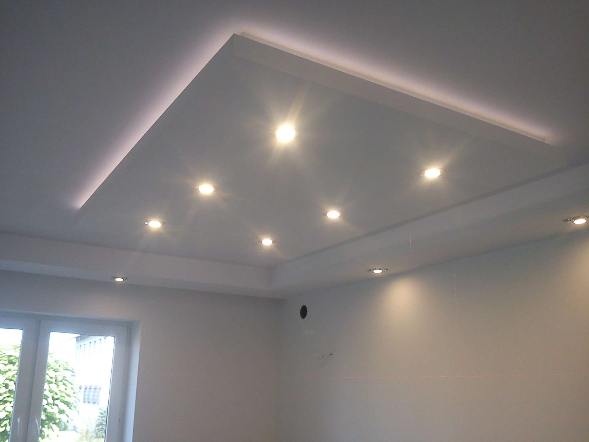 Biały sufit podwieszany z oświetleniem LED w kształcie prostokąta i punktowymi lampami halogenowymi, widok z dołu na fragment pokoju z oknem.