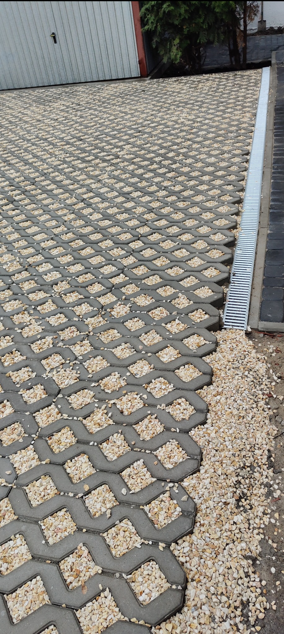 Ułożona szara ażurowa nawierzchnia z wypełnieniem z drobnych, jasnych kamyczków, obok odpływ liniowy.