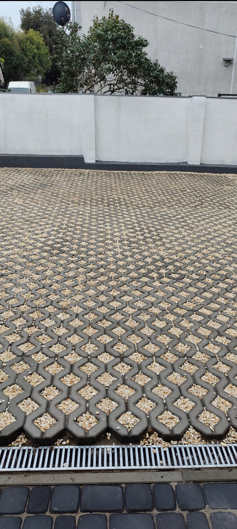 Ułożona nawierzchnia z ażurowych betonowych płyt wypełnionych drobnym kruszywem, z widocznym odpływem liniowym przy krawędzi, na tle białego muru i zieleni.