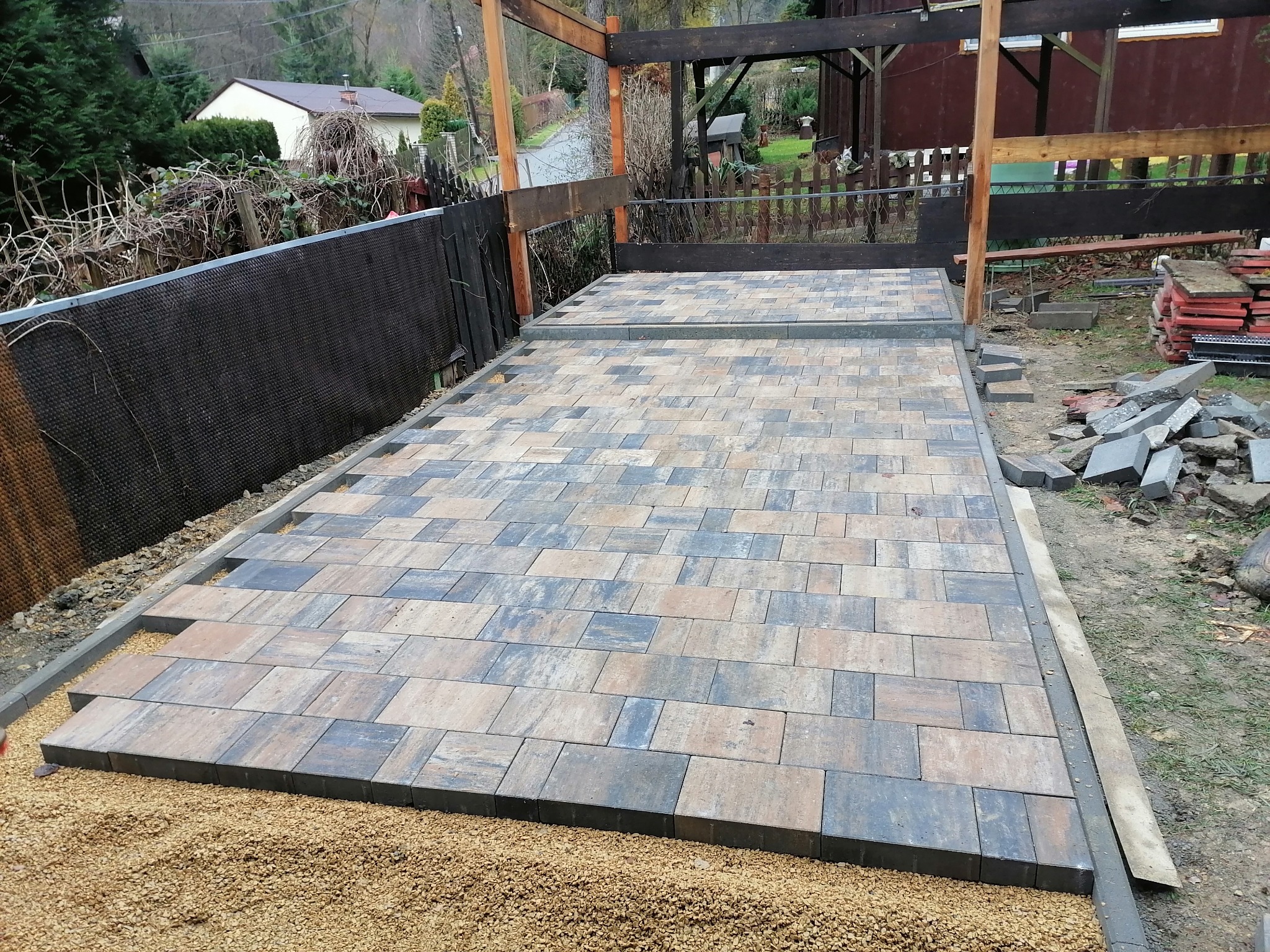 Fragment nowo ułożonej nawierzchni z kostki brukowej w odcieniach szarości i beżu, z widoczną podsypką żwirową i elementami konstrukcji z drewna w tle.