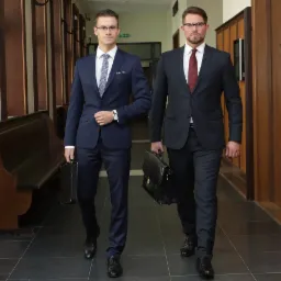 Dwóch elegancko ubranych mężczyzn w garniturach idzie korytarzem, trzymając aktówki. W tle widoczne drewniane panele i ławki.