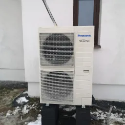 Zewnętrzna jednostka pompy ciepła Panasonic Inverter 9kW, zamontowana na czarnych bloczkach betonowych przy białej ścianie budynku w Płocku. Widoczne przewody wchodzące w ścianę oraz okno z brązową...