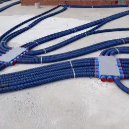 Niebieskie rury karbowane, połączone w rozdzielacze podłogowe z czerwonymi końcówkami, zamocowane metalowymi paskami do surowego betonowego podłoża.