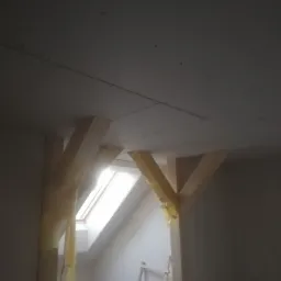 Poddasze w trakcie adaptacji: widoczne drewniane belki wzmacniające konstrukcję, zabezpieczone folią malarską, oraz zamontowana płyta kartonowo-gipsowa na suficie, z oknem dachowym w tle.