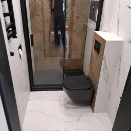 Nowoczesna łazienka z kabiną prysznicową z deseniem chevron na ścianie, wiszącą czarną toaletą i białymi płytkami imitującymi marmur na podłodze i ścianach.