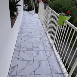 Białe płytki imitujące marmur ułożone na balkonie, widoczne białe balustrady, doniczki z kwiatami zawieszone na balustradach, w tle zieleń.