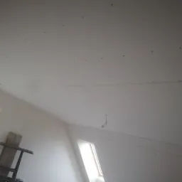 Skośny sufit na poddaszu pokryty płytami kartonowo-gipsowymi, widoczne ślady po wkrętach, okno dachowe i fragment drabiny malarskiej w rogu.