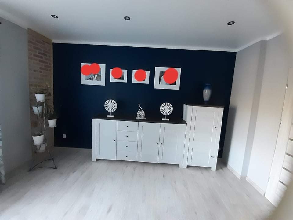 Jasna podłoga w salonie z ciemną ścianą, białymi meblami i dekoracjami ściennymi w białych ramkach, uzupełniona ceglaną ścianą z lewej strony i stojakiem na kwiaty.