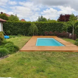 Niebieski basen otoczony drewnianym tarasem w ogrodzie z tujami w tle, widoczny zwinięty żółty wąż ogrodowy na trawie.