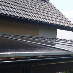 Zadaszenie tarasu wykonane z poliwęglanu, mocowane do ściany budynku z widocznym systemem odprowadzania wody deszczowej, z perspektywy z dołu.