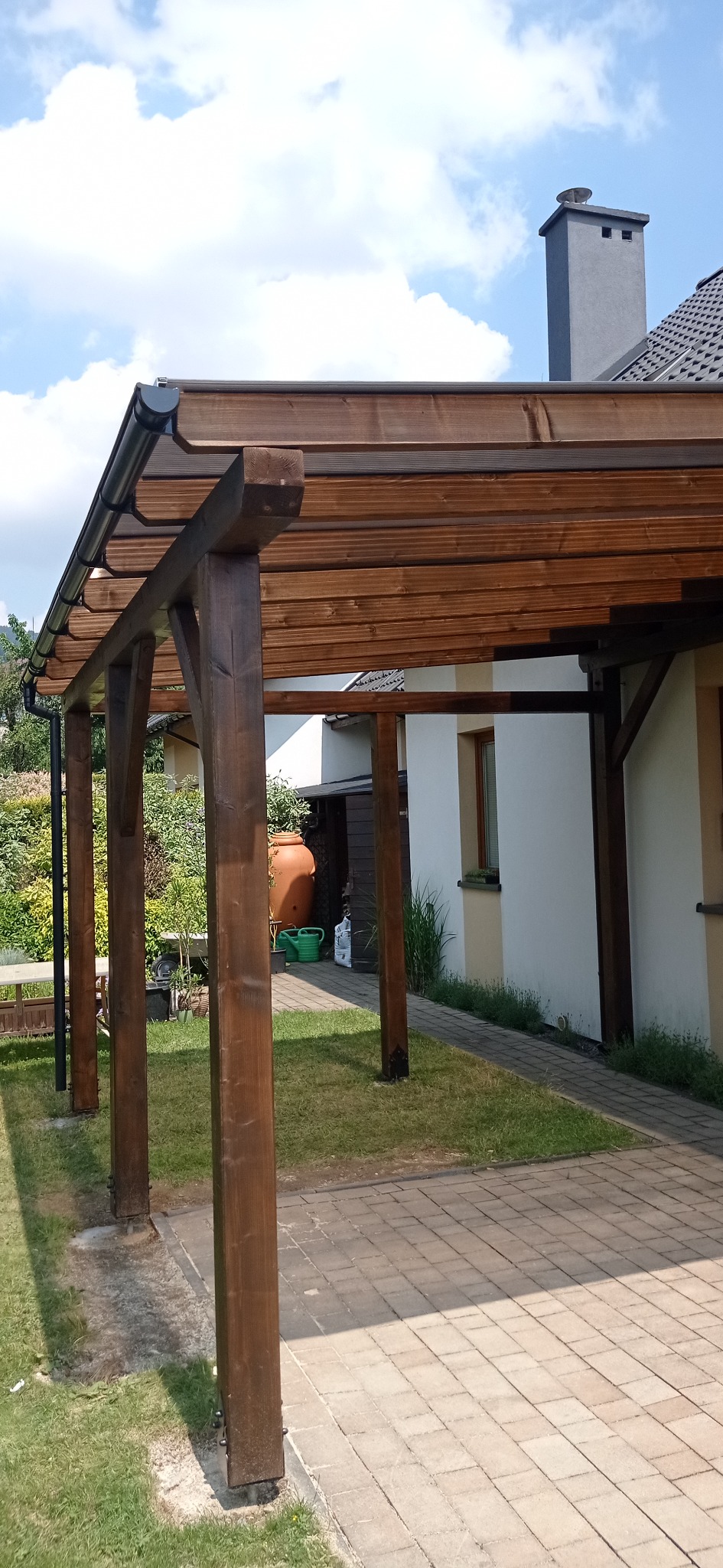 Zadaszona pergola z ciemnego drewna, z widocznymi elementami mocującymi i rynną, przylegająca do budynku z jasną elewacją i szarym kominem, na tle zieleni i błękitnego nieba.