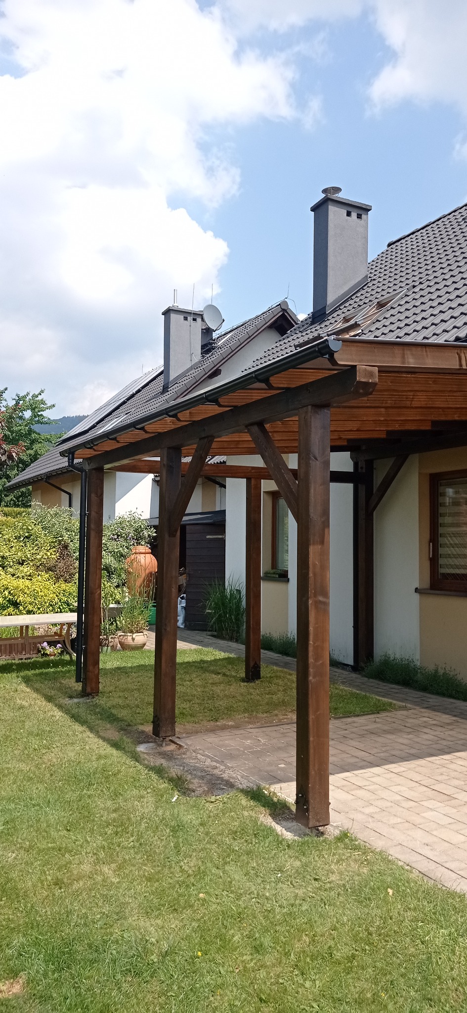 Drewniana konstrukcja z brązowymi słupami tworząca zadaszenie przy domu z szarą dachówką, widoczne panele słoneczne na dachu i zadbany trawnik
