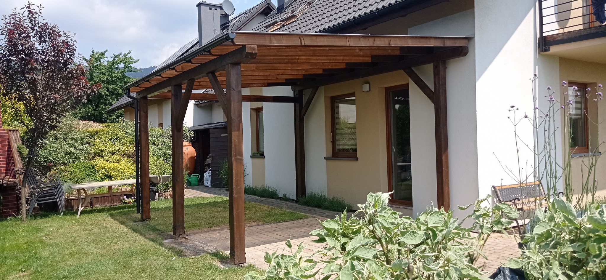 Drewniana konstrukcja zadaszenia przylegająca do domu z widocznymi elementami architektury ogrodowej, takimi jak roślinność i meble ogrodowe.